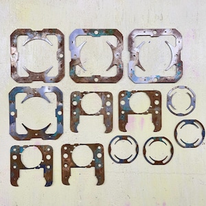 Puede incluir: Colección de juntas metálicas oxidadas de varias formas y tamaños. Las juntas tienen un marco exterior cuadrado o circular con recortes interiores. Algunas muestran rastros de pintura azul y verde, y todas tienen un aspecto desgastado.