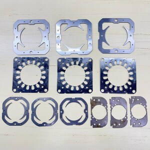 May include: A collection of various silver-colored metal gaskets and plates. The shapes include squares, rectangles, and partial circles with cut-out designs. Some have circular openings, while others have unique geometric patterns.