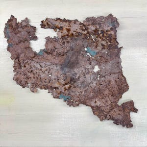 May include: A textured, abstract metal art piece with a rusty brown and teal color palette. The artwork has an irregular shape with a rough, weathered appearance, suggesting an industrial or reclaimed aesthetic. The piece is mounted on a light-colored wooden surface.