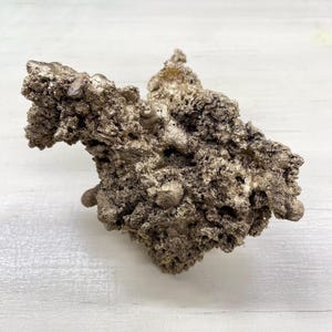 Könnte beinhalten: Silberfarbene Mineralprobe mit unregelmäßiger Form und rauer, strukturierter Oberfläche. Die Probe hat ein poröses Aussehen mit kleinen, abgerundeten Vorsprüngen. Die Gesamtform ist organisch und natürlich.