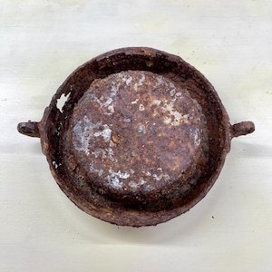 May include: A rusty, circular metal object with a raised rim and two small handles. The object has a weathered appearance with areas of corrosion and discoloration, suggesting age and exposure to the elements. The overall shape is reminiscent of a small pan or dish.