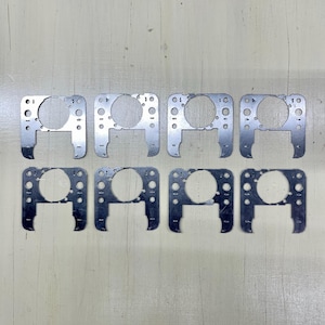 May include: Eight silver-colored metal brackets are arranged in two rows. Each bracket has a circular opening in the center, with multiple smaller holes around the perimeter. The brackets have a rectangular shape with a curved bottom.