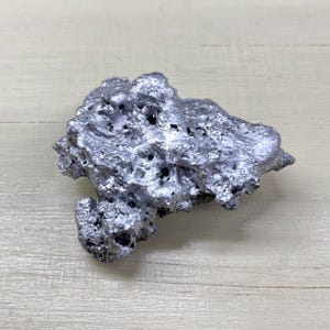 Könnte beinhalten: Ein silberfarbenes, unregelmäßig geformtes Objekt mit poröser Textur. Das Objekt hat eine raue, unebene Oberfläche mit zahlreichen kleinen Löchern und metallischem Glanz. Es liegt auf einer hellen Holzoberfläche.