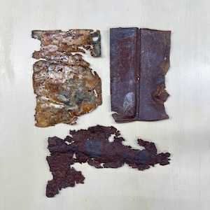 Peut inclure: Quatre morceaux de métal rouillé, présentant des formes et des textures variées. Les couleurs dominantes sont le marron et l'orange, avec des touches de vert, indiquant l'oxydation. Les pièces sont disposées sur une surface claire.