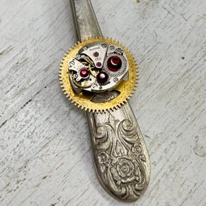 May include: A gold-toned gear with a silver watch movement inside. The watch movement has three red jewels and the text "HMT 17J" is visible.