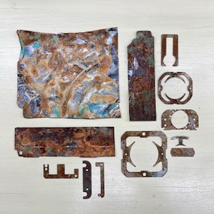 May include: An assortment of irregularly shaped, rusted metal pieces with shades of brown, copper, and teal. The collection includes a crumpled sheet, rectangular plates, and various cut-out shapes, all displaying signs of oxidation.