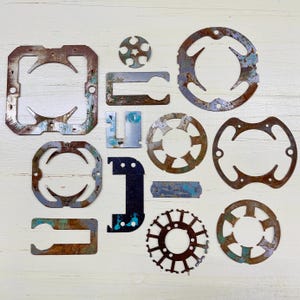 May include: An assortment of rusted metal cutouts in various shapes, including squares, circles, and brackets. The pieces have a distressed appearance with shades of brown, rust, and hints of blue-green. The objects are arranged on a white wooden surface.