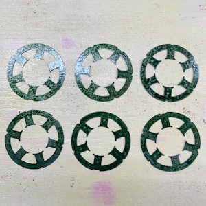 Puede incluir: Seis objetos circulares de metal verde oscuro con un patrón repetitivo de formas recortadas. Los objetos están dispuestos en dos filas de tres sobre una superficie de color claro. Las formas recortadas recuerdan a una estrella o una flor.