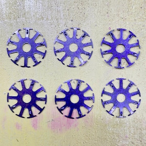 Puede incluir: Seis objetos circulares de color púrpura metálico con un agujero central y un diseño de radios. Los objetos están espaciados uniformemente sobre una superficie de color amarillo claro. Cada objeto tiene una serie de recortes espaciados uniformemente alrededor del perímetro.