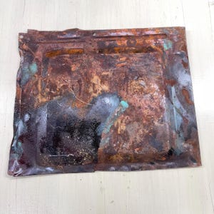 May include: A rectangular metal sheet with a heavily oxidized surface. The sheet displays a mix of brown, black, and teal colors, with visible signs of corrosion and wear. The edges are slightly bent, and there are a few small holes.