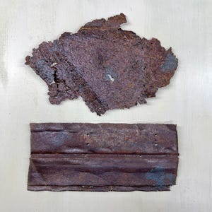 Peut inclure: Deux morceaux de métal rouillé et vieilli. La pièce supérieure est de forme irrégulière, ressemblant à un fragment déchiré. La pièce inférieure est rectangulaire avec plusieurs petits trous et un bord plié. Les deux pièces sont d'un brun rougeâtre profond.