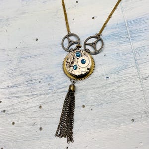 Puede incluir: Un collar de cadena dorada con un colgante de estilo steampunk que presenta una esfera de reloj circular con diamantes de imitación azules y engranajes. El colgante está unido a dos anillos plateados y un borla de cadena dorada.