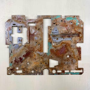 Peut inclure: Une pièce métallique rectangulaire et vieillie avec un design géométrique complexe. La surface présente un mélange de couleurs rouille-brun, turquoise et argent, avec de nombreux trous et formes découpées. L'objet a une esthétique industrielle et usée.