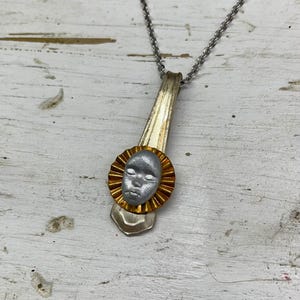 May include: A silver spoon necklace with a gold-rimmed pendant featuring a sculpted face. The pendant is a circular shape with a textured, sun-like design around the face.