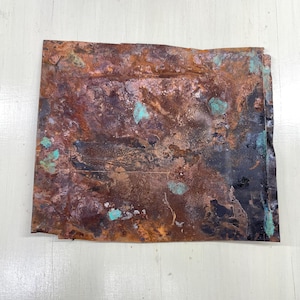 May include: A rectangular sheet of weathered copper with a rich, mottled appearance. The surface displays a blend of brown, rust, and black hues, accented by patches of turquoise-green oxidation. The edges are slightly uneven, adding to the aged aesthetic.