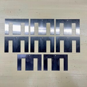 May include: A collection of eight metal plates, each with a unique U-shaped cutout. The plates are arranged on a light-colored wooden surface. Each plate has a small hole near the top.