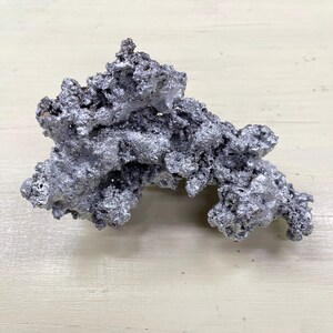 Könnte beinhalten: Ein silberfarbenes, unregelmäßig geformtes Objekt mit rauer, strukturierter Oberfläche. Das Objekt hat ein poröses Aussehen mit zahlreichen kleinen Löchern und Spalten. Es liegt auf einer hellen Holzoberfläche.