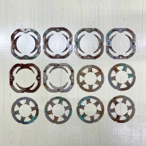 May include: A collection of twelve weathered metal decorative pieces. The pieces are in various shapes, including squares and circles, with cut-out designs and a distressed, aged appearance. The metal has a mix of brown, gray, and teal colors.