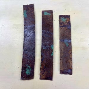 Peut inclure: Trois morceaux rectangulaires de métal vieilli, de couleur brun rouille avec des taches d'oxydation verte. Les bandes métalliques, de longueurs et largeurs variables, ont des bords rugueux et irréguliers.