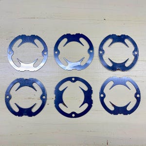 May include: Six circular metal pieces with a shiny, reflective surface. Each piece has a central opening and a unique, curved design with three points extending inward. Small holes are evenly spaced around the outer edge of each circle.