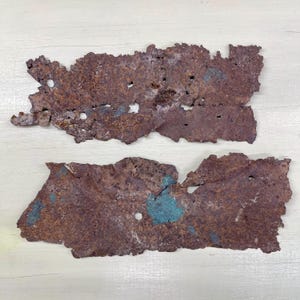May include: Two irregularly shaped pieces of rusted metal with a dark brown color and areas of turquoise-green oxidation. The metal has a rough, textured surface with several small holes and jagged edges. The pieces are displayed on a light-colored wooden surface.