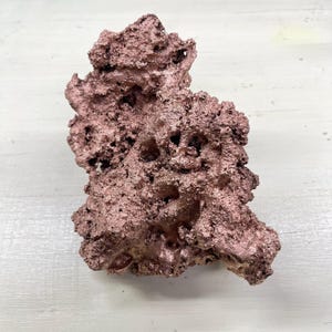 Könnte beinhalten: Ein rohes, unregelmäßig geformtes Kupferstück mit einer strukturierten, porösen Oberfläche. Das Kupfer hat eine rotbraune Farbe und scheint unpoliert zu sein. Das Stück wird vor einem weißen Hintergrund präsentiert, um seine natürliche Form und seinen metallischen Glanz hervorzuheben.
