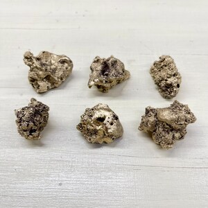 Könnte beinhalten: Sechs goldfarbene Nuggets unterschiedlicher Größe und Textur sind auf einer weißen Holzoberfläche angeordnet. Die Nuggets haben ein raues, poröses Aussehen, wobei einige dunklere, möglicherweise oxidierte Bereiche aufweisen. Die Beleuchtung hebt den metallischen Glanz des Goldes hervor.