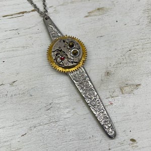 Puede incluir: Un collar de plata con un largo colgante grabado y una esfera de reloj dorada con engranajes. La esfera del reloj tiene una joya roja en el centro.