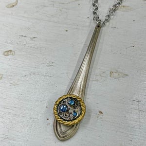 以下が含まれることがあります： 銀のスプーンネックレス。ゴールドの縁取りが施された円形のペンダントには、青いラインストーンと歯車が付いたスチームパンクのデザインが施されています。