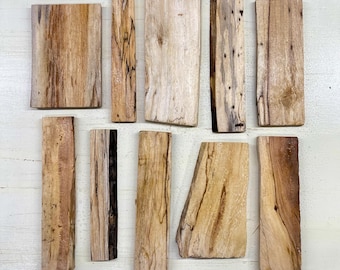May include: A collection of eleven irregularly shaped wooden planks. The planks vary in size and shape, with natural wood grain patterns in shades of light brown, tan, and darker brown. Some planks have a mix of colors, including black and gray.