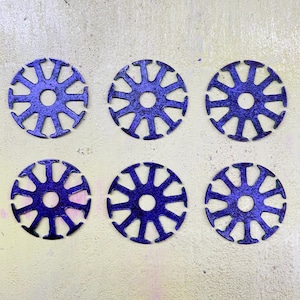 Puede incluir: Seis elementos decorativos circulares azules con un diseño de estrella. Cada elemento tiene un agujero central y brazos radiales, creando un patrón simétrico. Los objetos están dispuestos sobre una superficie de color claro.