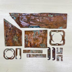 May include: An assortment of irregularly shaped copper pieces with a weathered, aged appearance. The copper displays a mix of brown, orange, and green hues, suggesting oxidation. The collection includes rectangular, triangular, and uniquely cut shapes, possibly for artistic or industrial use.