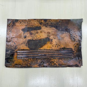 May include: A rectangular piece of metal with a distressed, aged appearance. The metal has a mix of copper, brown, and black tones, with areas of oxidation and texture. Horizontal lines and a circular element are visible.