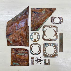 May include: An assortment of irregularly shaped, weathered copper pieces with a mix of brown, green, and blue-green patinas. The collection includes various geometric shapes, including squares, circles, and angular fragments, suggesting industrial salvage or repurposed materials.