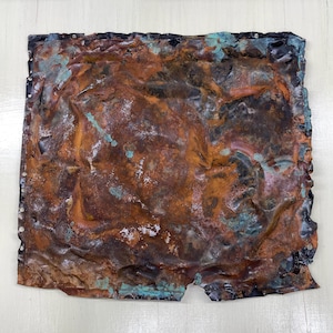 Könnte beinhalten: Ein quadratisches Metallkunstwerk mit einem verwitterten, gealterten Aussehen. Das Stück weist eine Mischung aus rostfarbenen, orangefarbenen und türkisfarbenen Farbtönen auf, mit sichtbaren Gebrauchsspuren. Die Kanten sind uneben, und kleine Nieten sind sichtbar.