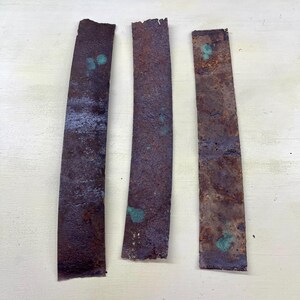 Peut inclure: Trois bandes de métal rectangulaires rouillées, de couleur brun foncé. Des taches d'oxydation bleu-vert sont visibles. Les bords sont irréguliers et les largeurs variables, suggérant une récupération ou une réutilisation.