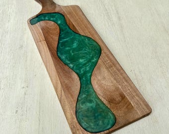 May include: Wooden serving board with a handle, featuring a river of green epoxy resin. The board is rectangular with a carved-out section filled with the swirling green resin. The wood has a natural grain.