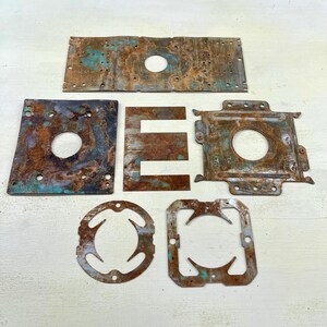 Peut inclure: Ensemble de sept pièces métalliques rouillées et patinées, de formes variées, dont des rectangles, des carrés et une lettre "E". Le métal présente des zones de patine bleu-vert, indiquant l'âge et l'exposition. Certaines pièces ont des trous.