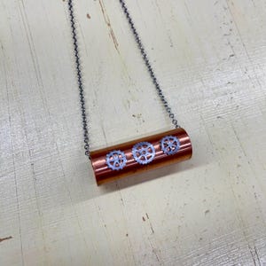 Puede incluir: Un collar de estilo steampunk con un colgante cilíndrico de color cobre adornado con tres diseños de engranajes plateados. El colgante cuelga de una delicada cadena plateada. El collar tiene una estética industrial única.