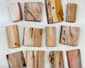 May include: A collection of twelve irregularly shaped wooden blocks. The blocks vary in size and shape, with natural wood grain patterns in shades of tan, brown, and pink. Some blocks have bark edges.