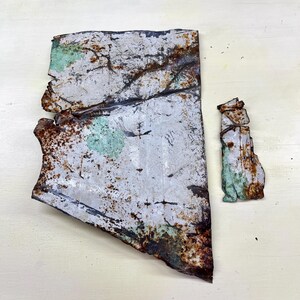 Peut inclure: Deux morceaux de métal vieilli à l'aspect usé. La plus grande pièce est de forme irrégulière, principalement blanche avec des zones de rouille et de vert. La plus petite pièce est un rectangle vertical avec une coloration et une texture similaires.