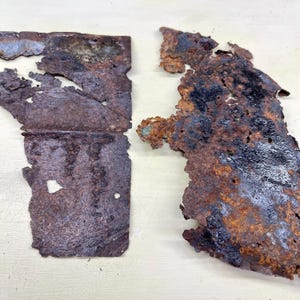 May include: Two pieces of heavily rusted metal with jagged edges. The metal is primarily brown with areas of black and hints of green. The pieces appear to have been torn or broken, revealing a rough texture.