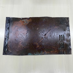 May include: A rectangular, weathered metal sheet with a dark brown and copper patina. The metal has a textured surface with three sets of three rectangular cutouts on the right side. The left edge has a black, riveted border.