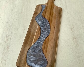 May include: Wooden serving board with a handle, featuring a river of swirling gray resin. The board is rectangular with a natural wood grain finish. The resin river design adds a decorative element to the serving board.