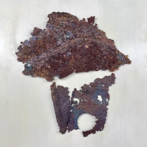 May include: Two irregularly shaped pieces of weathered metal with a textured surface. The top piece is primarily brown with hints of blue and purple. The bottom piece is a darker purple with a hole and a similar texture.
