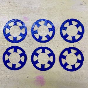 Puede incluir: Seis posavasos circulares de fieltro azul con un diseño simétrico calado. Cada posavasos presenta un círculo central rodeado por un anillo con recortes espaciados uniformemente, creando un efecto de estrella. Los posavasos están dispuestos sobre una superficie clara.