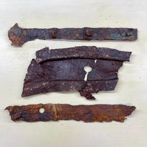 Peut inclure: Trois pièces de métal fortement rouillé, probablement d'un artefact historique. La pièce supérieure est une longue bande étroite avec des rivets. La pièce du milieu est un fragment plus grand, de forme irrégulière, avec un trou de serrure. La pièce inférieure est une bande étroite avec un trou.