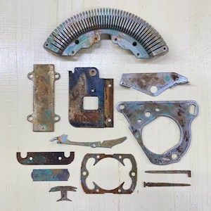 May include: An assortment of aged, weathered metal components with a patina of rust and verdigris. The collection includes a curved, slotted piece, brackets, and various shaped plates. The pieces are arranged on a light-colored surface, showcasing their aged appearance.