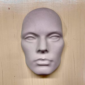May include: A pale lavender-colored, human-like face mask with closed eyes, a defined nose, and full lips. The mask is smooth and appears to be made of plaster or a similar material. The background is a light, textured surface.