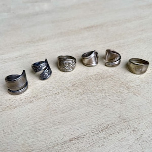 May include: Six silver rings with unique designs are arranged on a light-colored wooden surface. The rings vary in style, including a spiral design, floral details, and simple bands, showcasing a handcrafted jewelry collection.